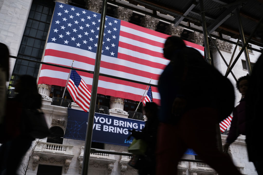 pessoas andando em frente à tela com bandeira dos Estados Unidos sendo projetada no mercado de ações de Nova York no contexto do ultimo pico de desemprego dos eua, a crise do coronavírus.