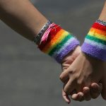 Duas mãos femininas segurando uma a outra e usando braceletes com cores da bandeira LGBT