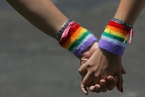 Duas mãos femininas segurando uma a outra e usando braceletes com cores da bandeira LGBT