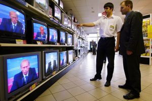 homem em frente a sessão de televisões numa loja assistindo enquanto todas as TVs transmitem um discurso de Vladimir Putin