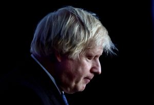 Boris Johnson de cabelos e rosto abaixado em semblante cansado e triste num fundo escuro